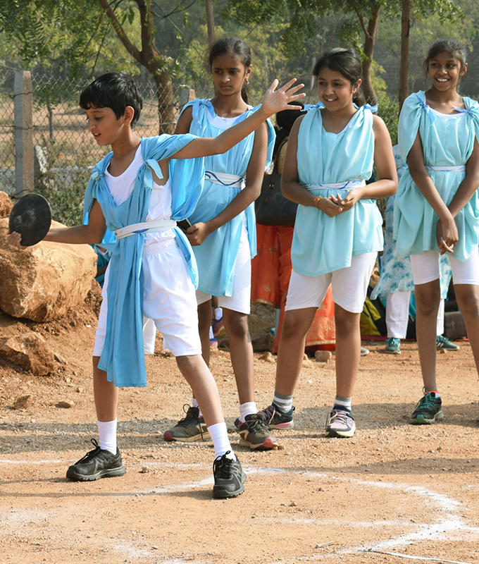 A middle school student dressed in traditional Greek garb engages in discus throw on a playground while classmates look on.