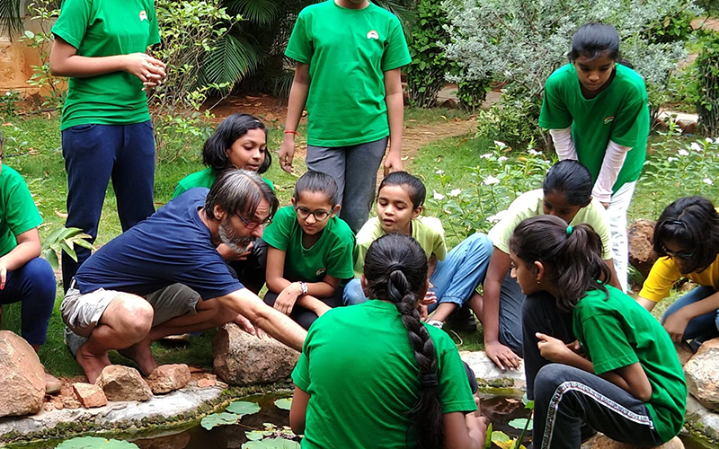 A group of students cluster around a pond in the garden at the Sloka campus, while a teacher points out something to them.