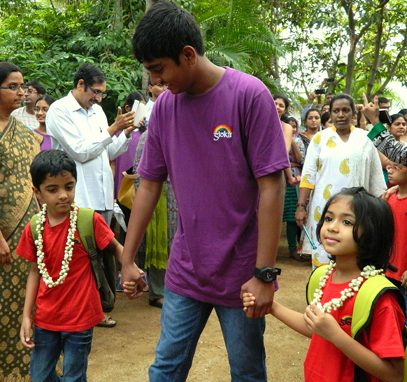 An older student holds the hands of two younger students on either side of him, outdoors surrounded by parents and teachers.