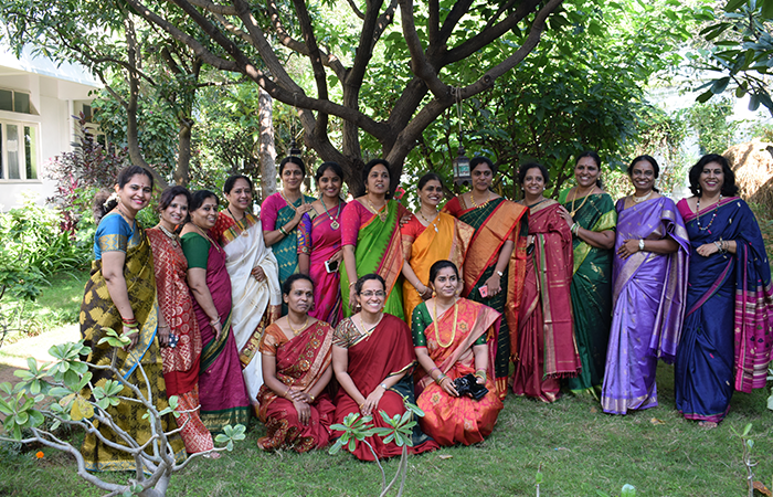 Several teachers dressed in colourful sarees pose facing the frame on a lawn in a garden at the Sloka campus.