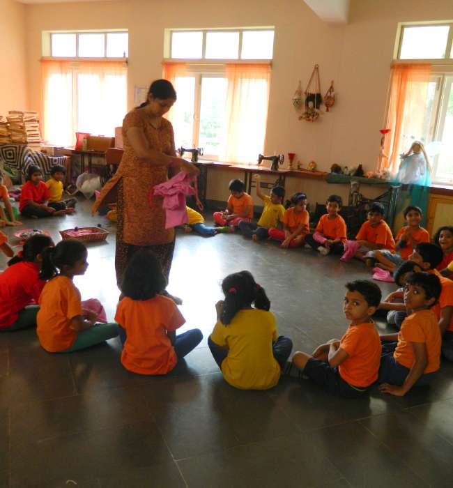 A group of young children sit in a circle on the classroom floor, two teachers stand beside, guide them in activity.