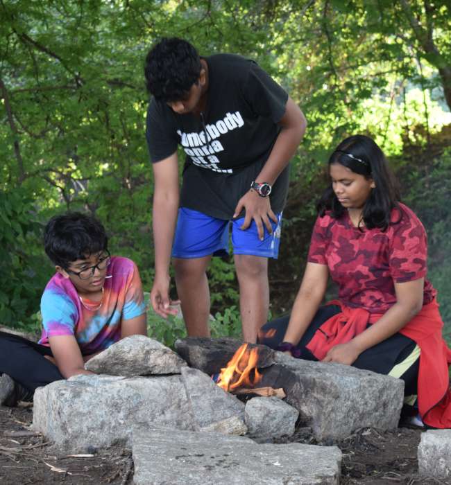 Three students tending to a fire burning within a stone stove during outdoor activity.