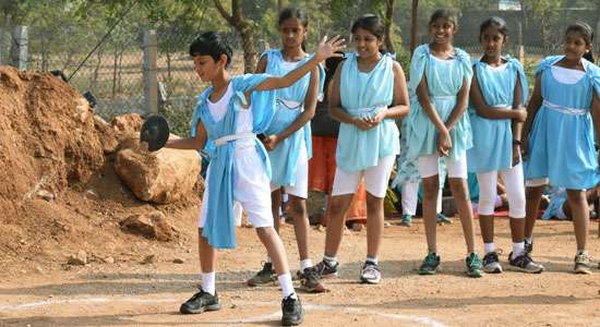 A middle school student dressed in traditional Greek garb engages in discus throw on a playground while classmates look on.