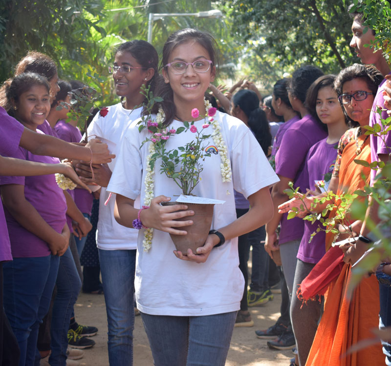 Students in white shirts carry flower pots in a row while students in purple shirts line the path on either side.