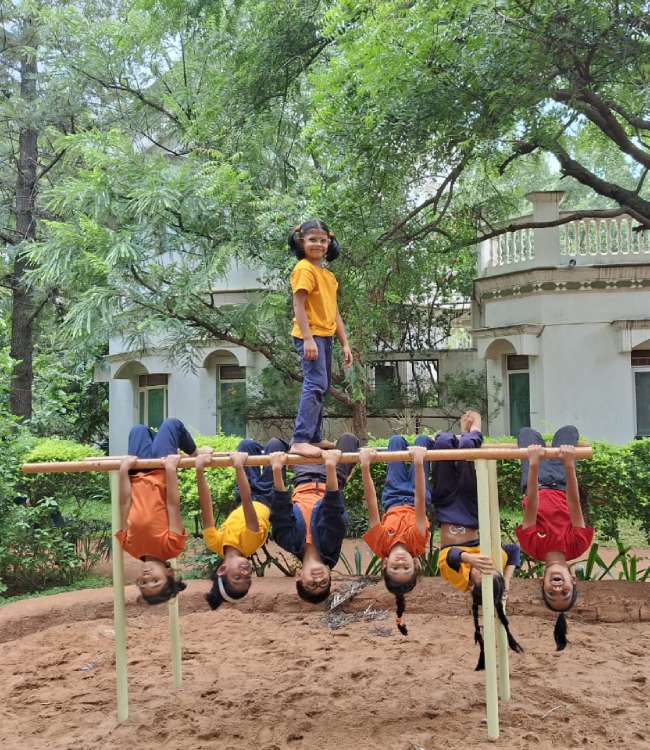 A student stands on a horizontal bar while six others hang upside down from it in an outdoor play area.