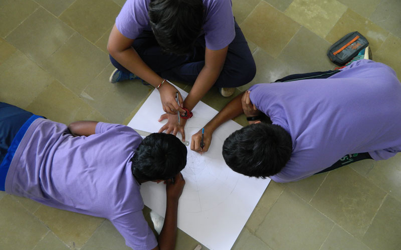 Three students wearing purple shirts bend over a paper sprawled on a floor, drawing a mandala.