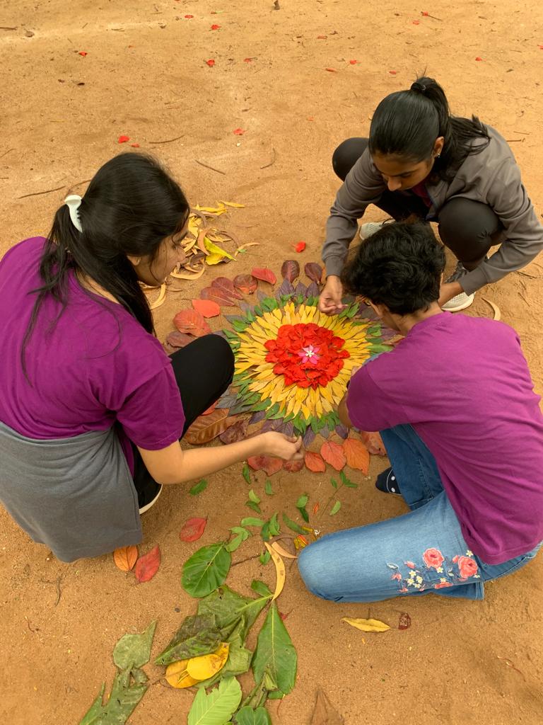 Three students squat on the ground making a colourful mandala from leaves and flowers.