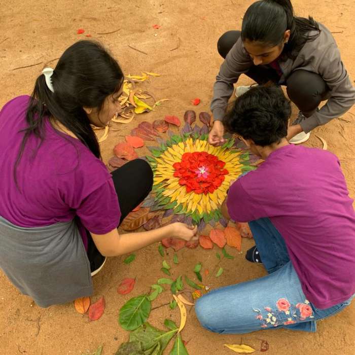 Three students squat on the ground making a vibrant mandala out of leaves and flowers.