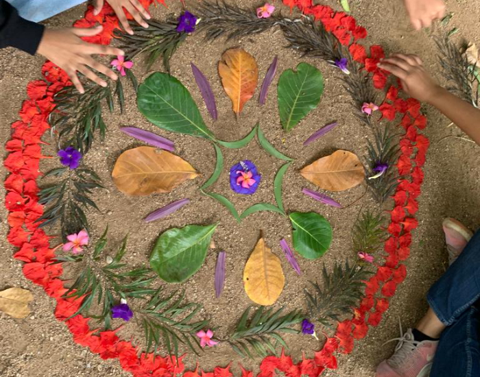 A couple of students hands are seen arranging a mandala on the ground, made of flowers and leaves