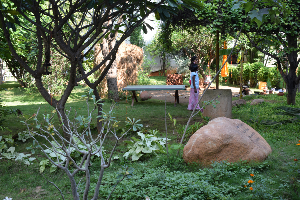 Two small children stand by a bench in a garden that contains a variety of plants, a rock etc.