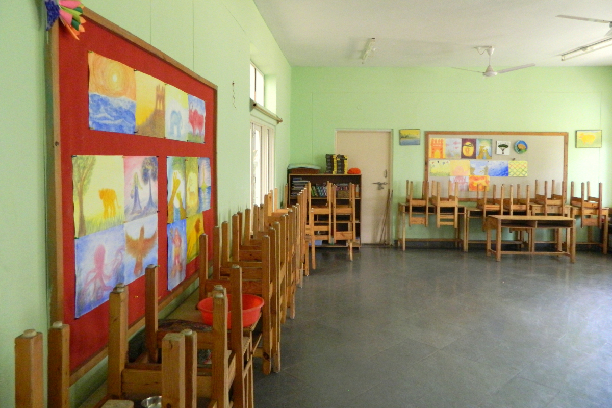 View of an empty kindergarten classroom with desks and chairs moved back against the wall; kids’ artworks line the walls.