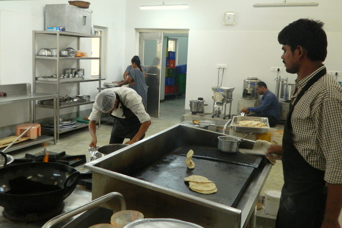 View of the school kitchen featuring a cook making chapatis on a stove and four other workers engaged in various activities.