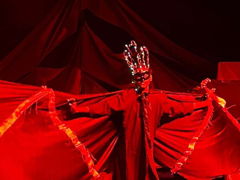 A student performs wearing a mask and costume, on a dark stage with red lighting.