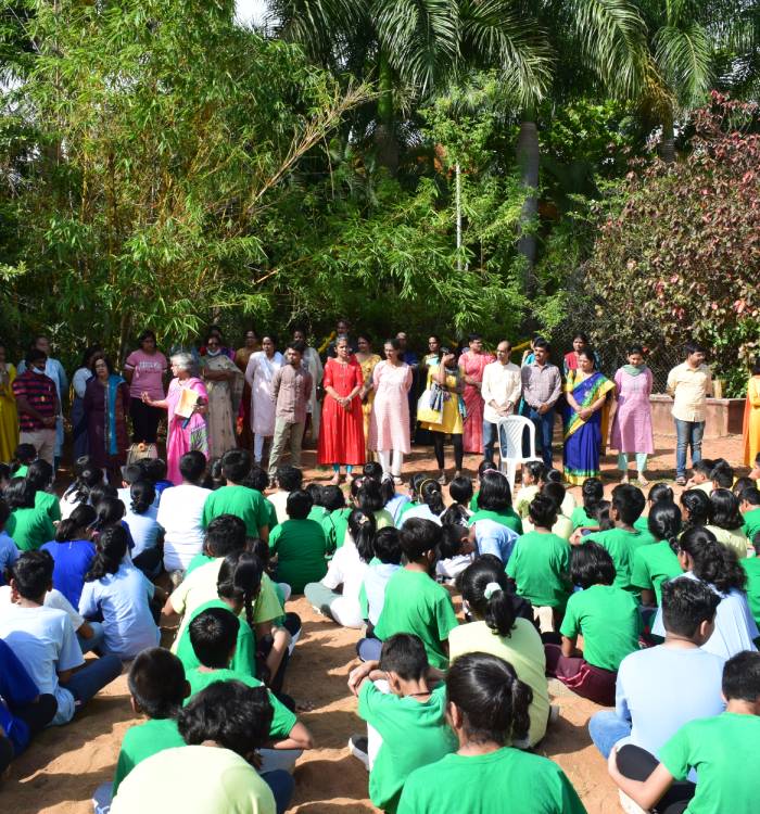 School assembly in progression; several students sit on the ground while teachers stand in a row in front of them.