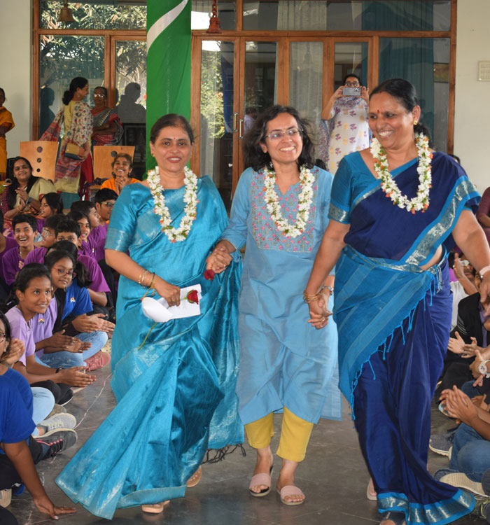 Three teachers wearing garlands walk together in a hall filled with seated students on either side during Rose Ceremony.